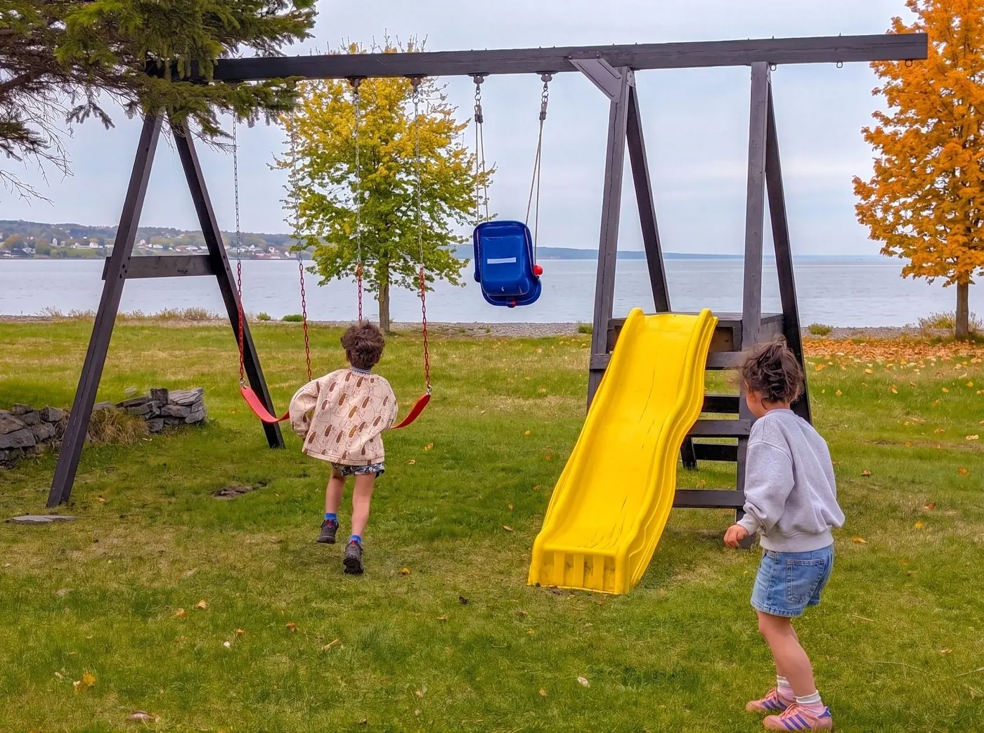 Enfants qui courent vers les balançoires et la glissade du parc à jeu, face au fleuve